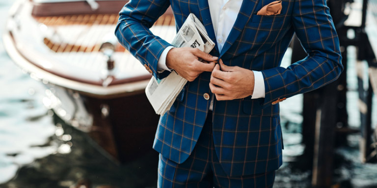 Man in an aquamarine blue suit with rust check and rust pocket square buttoning up his jacket while holding a newspaper in his right palm.
