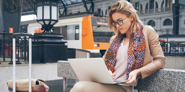 Young lady wearing glasses and a spotted scarf sitting at a train station working on her laptop.