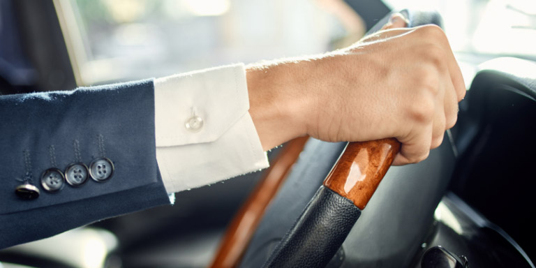 Man wearing a white shirt and blue suit jacket with his right hand on a wooden steering wheel.